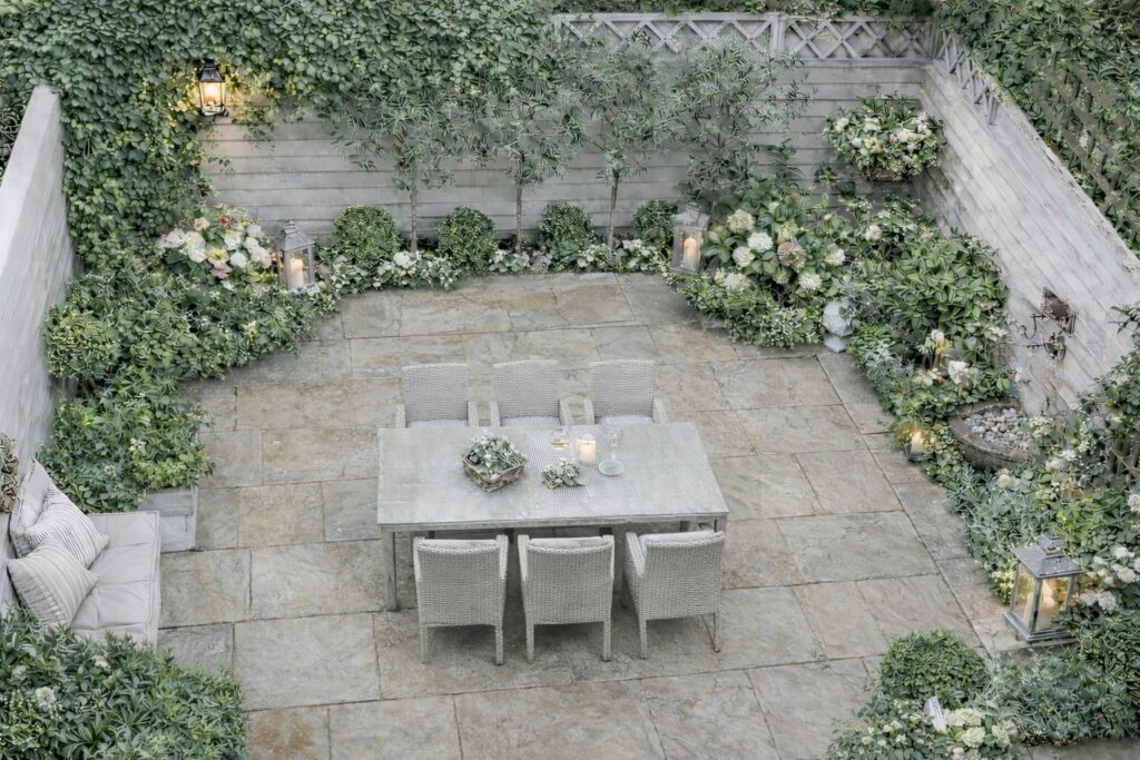 Un petit jardin paysagé en patio, clos, comprenant un ensemble de table à manger entouré d'une végétation luxuriante, incluant des arbustes, des plantes en pot et de petits arbres. Cet espace aménagé par un paysagiste dispose d'un sol en pierre claire et est entouré de clôtures en bois. L'entreprise de paysagisme a conçu cette prestation paysagère pour créer une ambiance apaisante et harmonieuse.